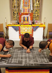 Chung Dolma (left), Sonam Dolma (middle), and Sherab Dolma (right) meticulously measure out and draw the geometric patterns of the mandala using compasses and rulers, with a colorful altar honoring the Buddha behind them